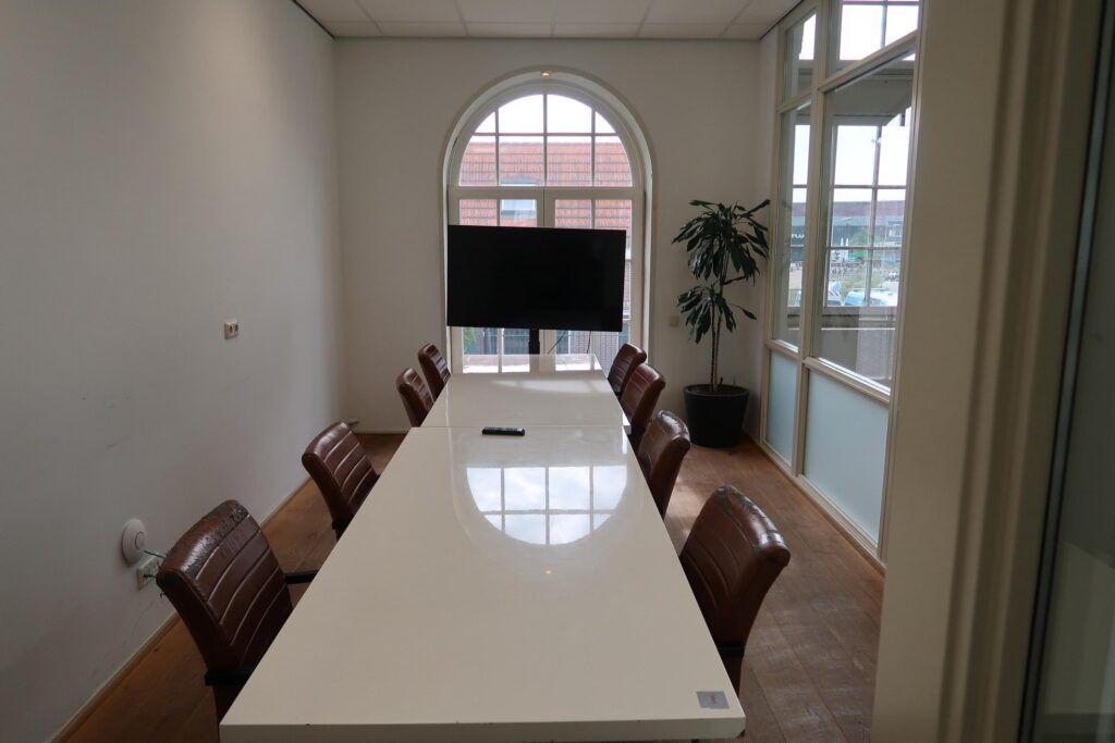 Vergaderruimte aan het Gedempt Hamerkanaal met lange witte tafel, leren stoelen, televisie en groot boograam.